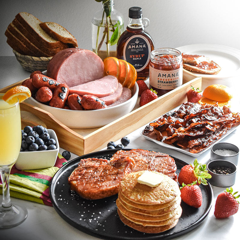 shot of brunch pack with pancakes and pork chops on a black plate the foreground; bacon, ham, sausages, syrup, and preserves in the background