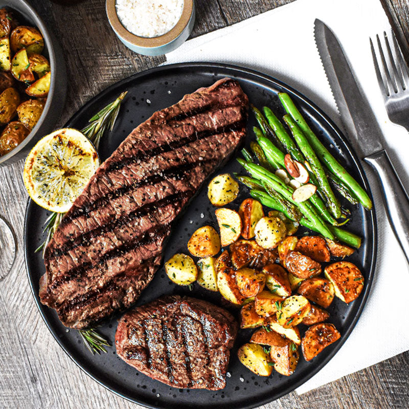 premium steak combo with potatoes and beans on a black tray along with silverware and glass on tabletop