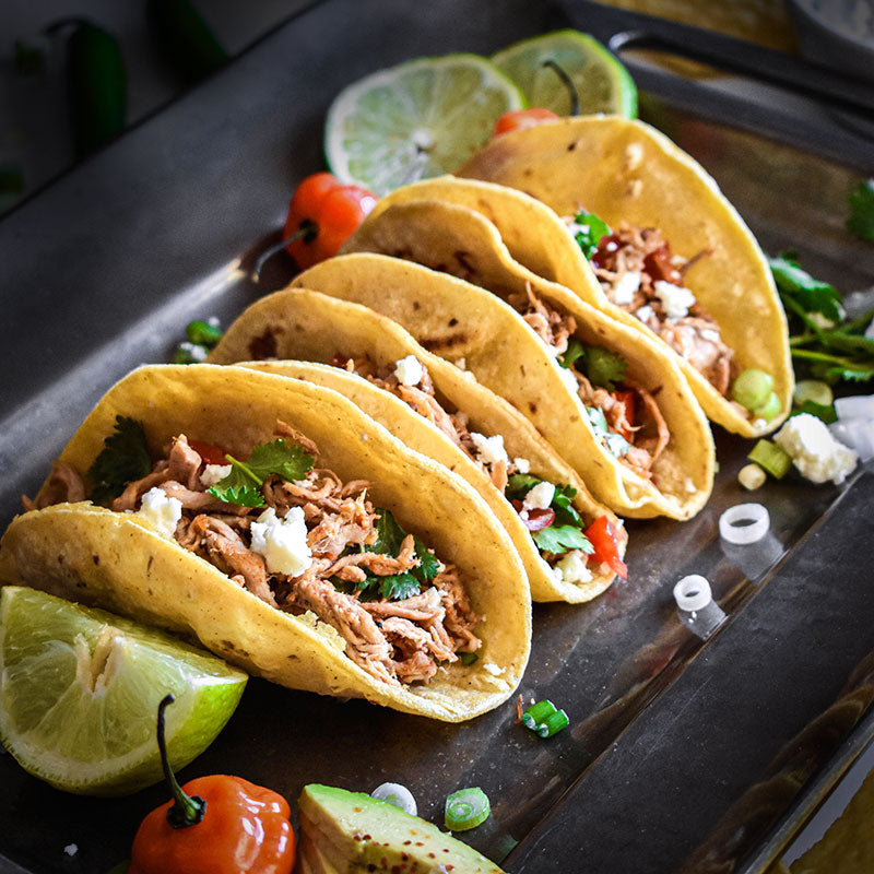 Shredded pork tacos on a tray with limes and peppers.