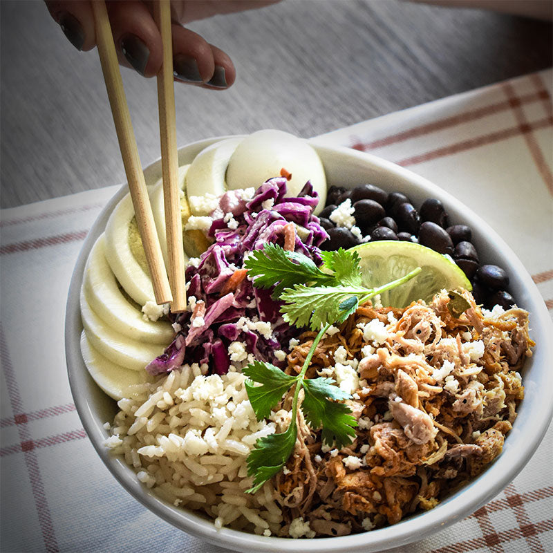 Rice bowl with shredded pork, beans, hard boiled egg, cabbage, and a hand using chopsticks to pick up food.