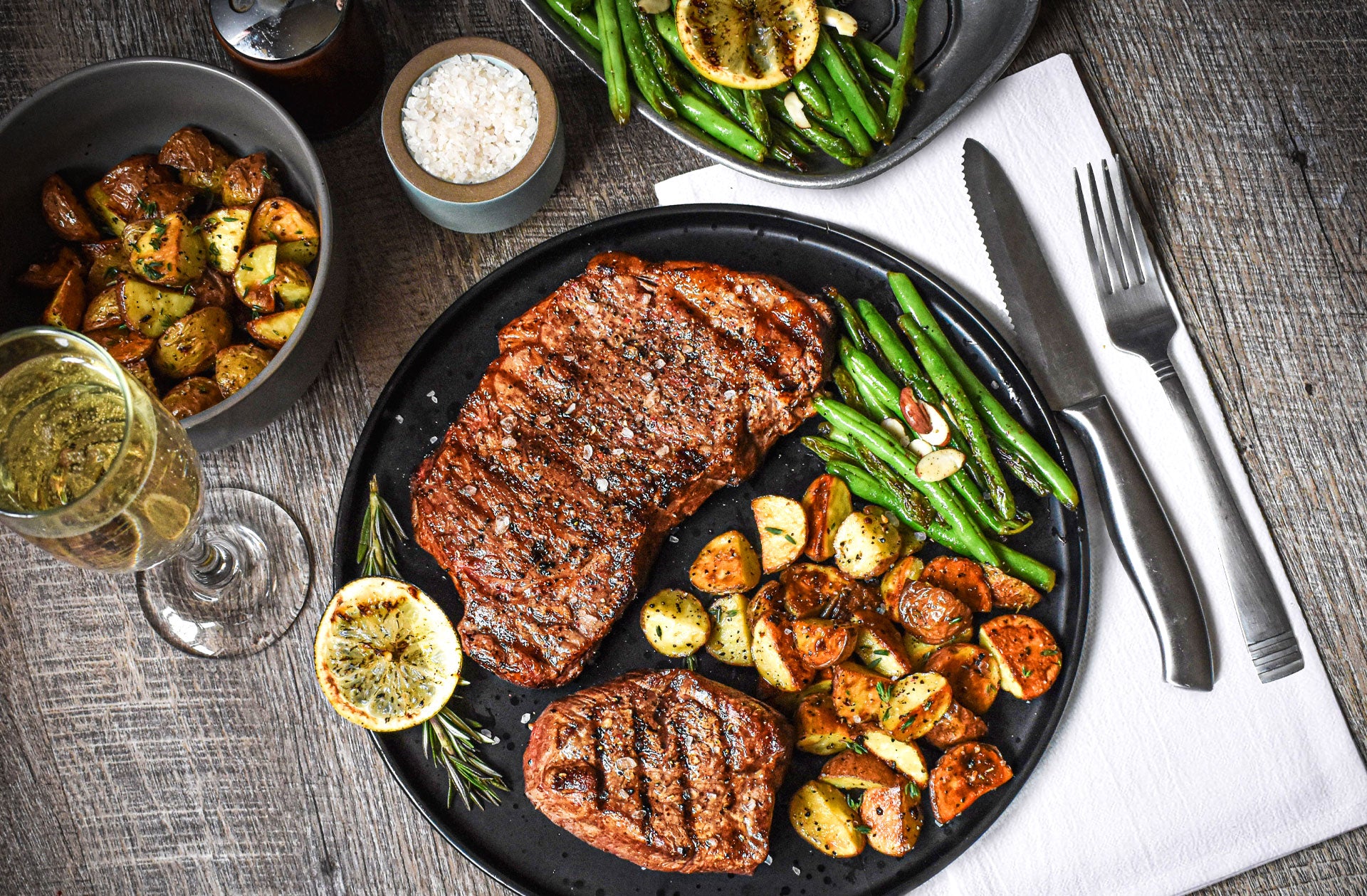 premium steak combo with potatoes and beans on a black tray along with silverware and glass on tabletop