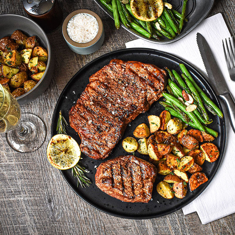 premium steak combo with potatoes and beans on a black tray along with silverware and glass on tabletop